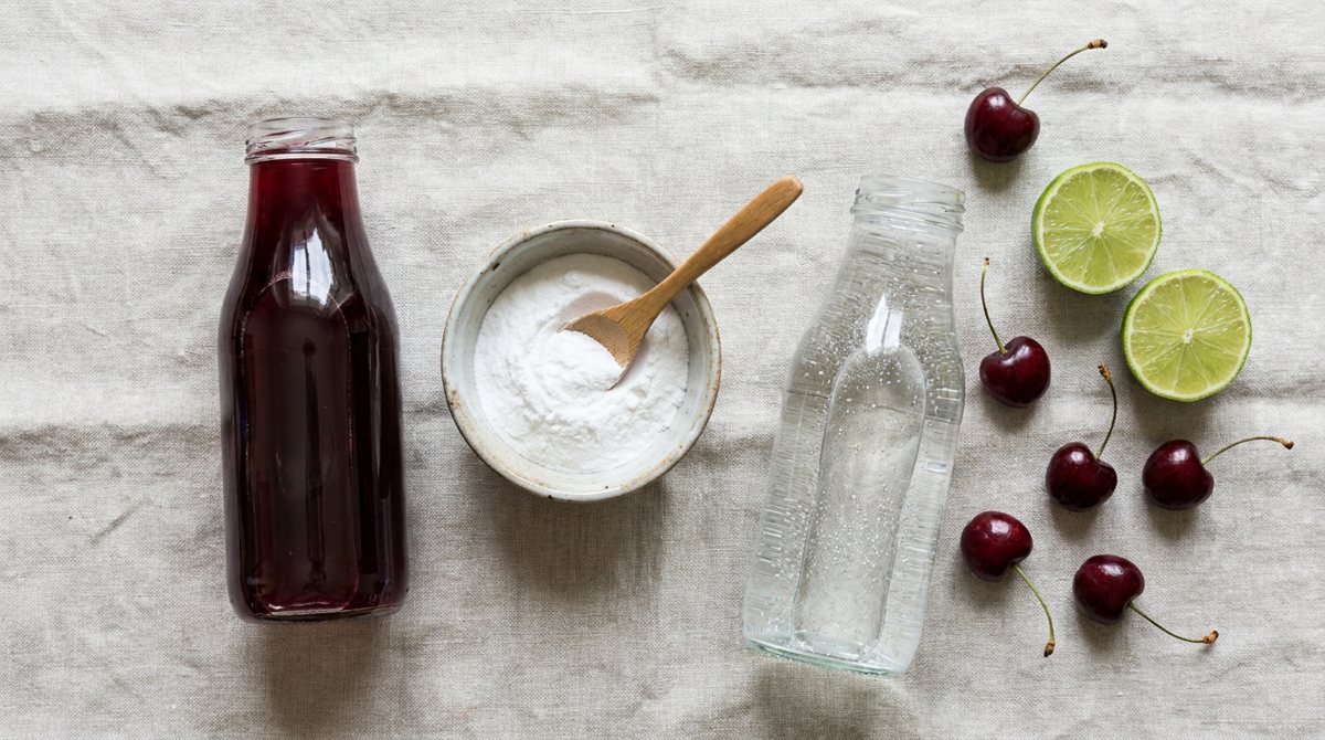 Flat lay of dark red cherry juice, a bowl of white powder, sparkling water, lime halves and whole cherries on linen